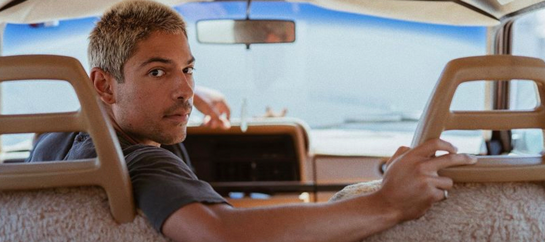 Person with short hair sitting in the backseat of a vehicle, looking back over their shoulder, with a hand resting on the seat.