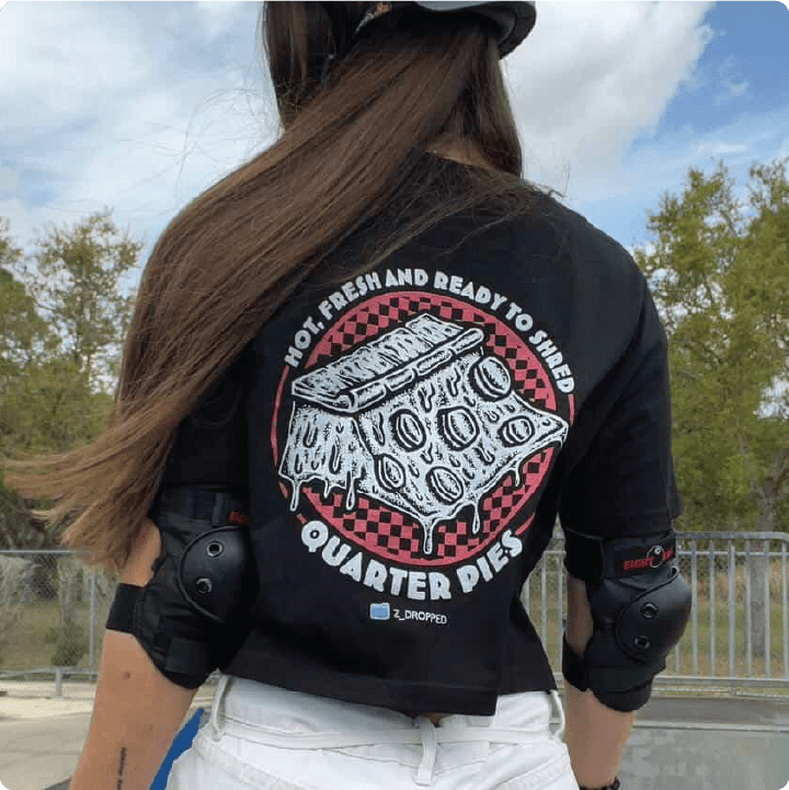 Rear view of a young skater girl wearing a black crop top shirt with a pizza quarter pipe graphic and text, at a skate park.