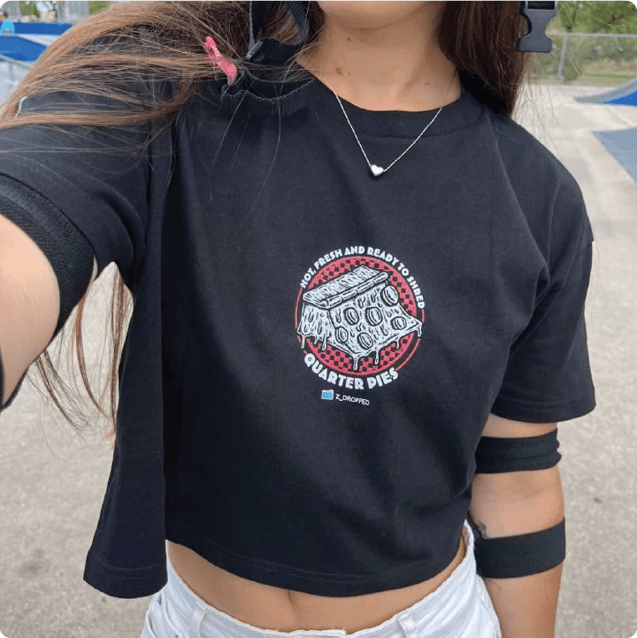 Young skater woman wearing a black crop top shirt with a pizza quarter pipe graphic and text, at a skate park.