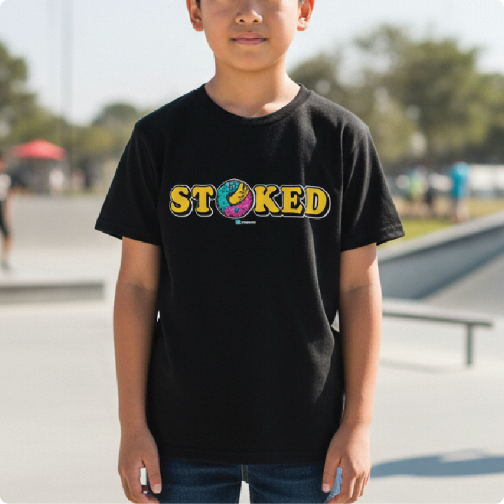 A child wearing a black short-sleeve t-shirt with a colorful 'STOKED' graphic across the chest, standing outdoors in a skatepark setting.