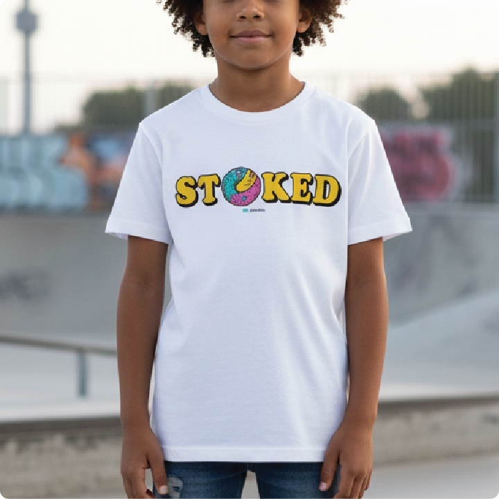 A child wearing a white short-sleeve t-shirt with a colorful graphic that reads 'STOKED' across the chest, standing in a outdoor skatepark setting.