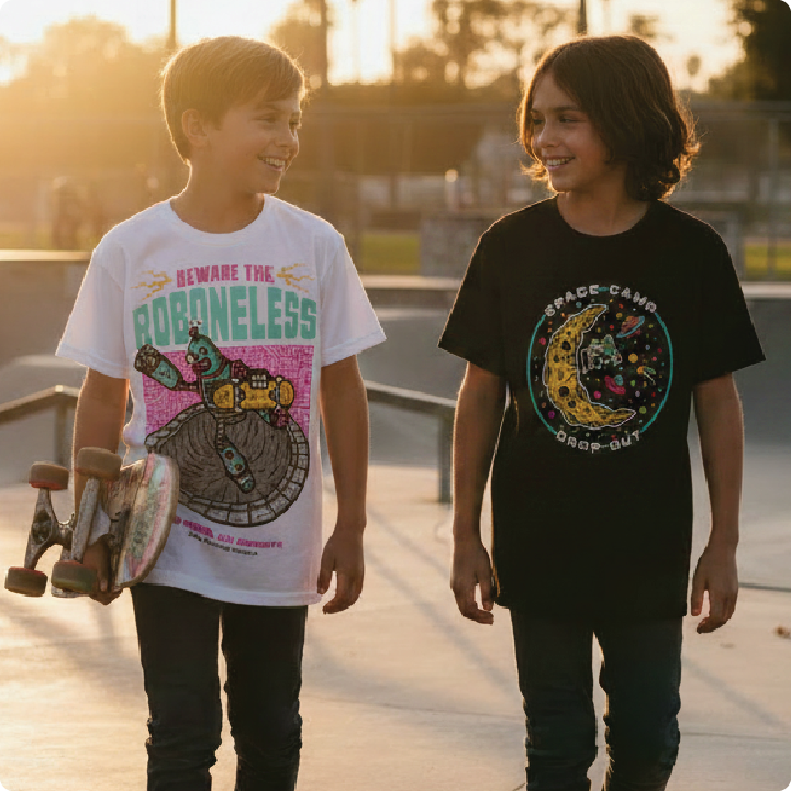 Two young kids walking with skateboards, wearing graphic t-shirts at a skate park.