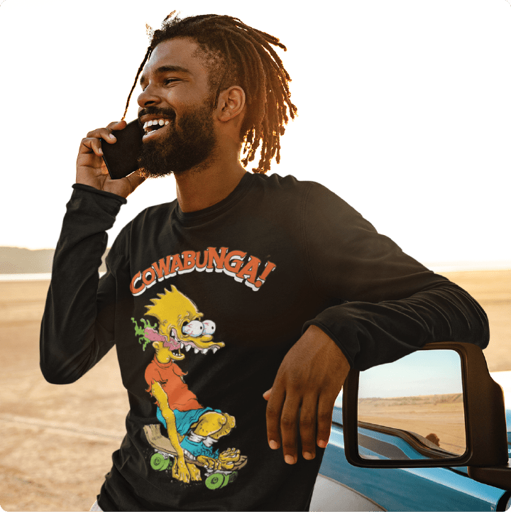 Young man wearing a black unisex zdropped Bart Roth long sleeve t-shirt on the phone in the desert by his car