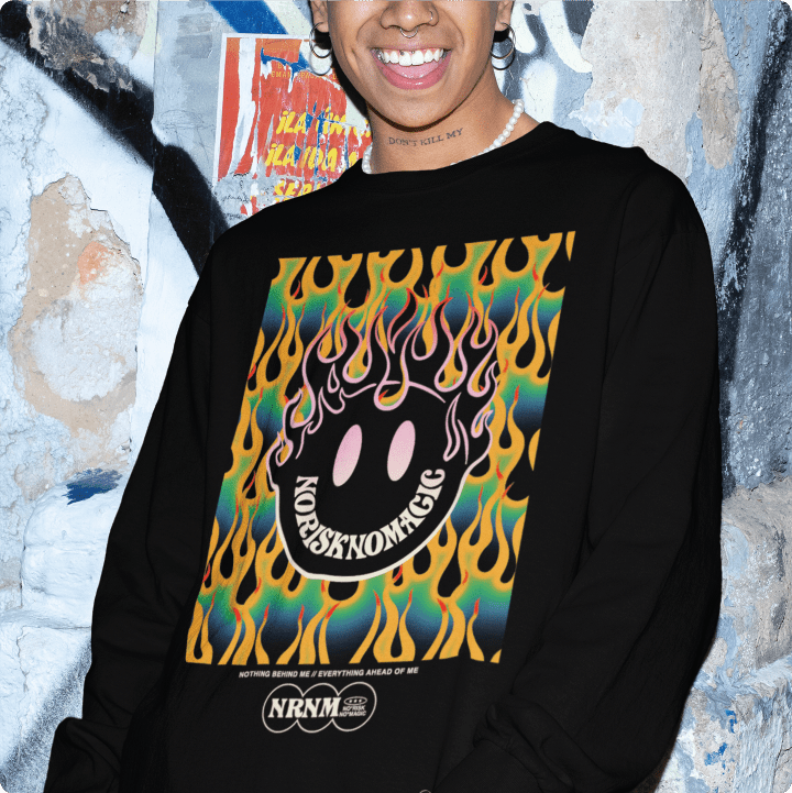 Person wearing a black long sleeve shirt with colorful flame smiley face design, standing against a graffiti-covered wall.