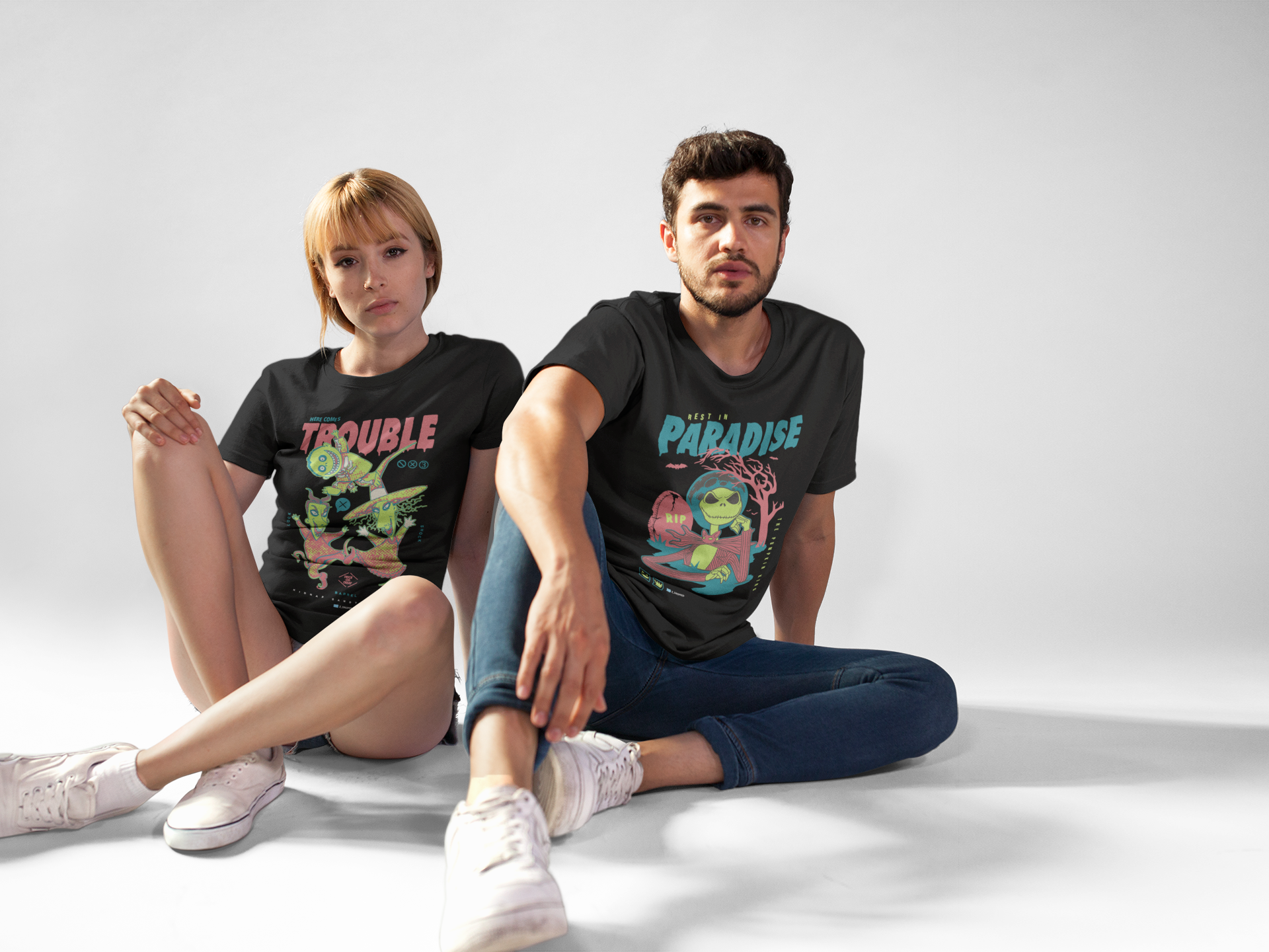Two people sitting on a white floor wearing colorful zdropped 'here comes trouble' graphic t-shirts.