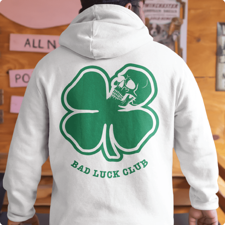Person wearing a white pullover hoodie with a green 4 leaf clover with a skull on the back, and 'Bad Luck Club' text at a bar setting.