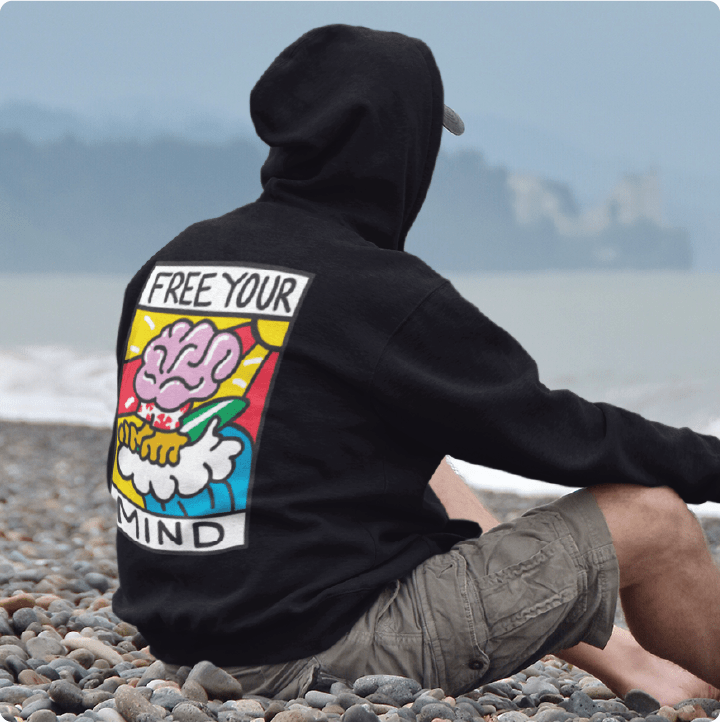 Person wearing a unisex black hoodie with Free Your Mind text and a brain surfing a wave on a pebbly beach.