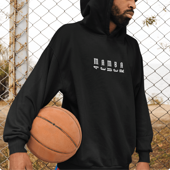 Young man wearing a black hoodie with white 'Mamba 4ever' text holding a basketball in front of a chain-link fence.