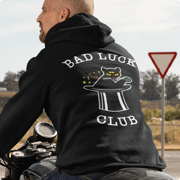 Rear view of a unisex black hoodie with white 'Bad Luck Club' text on the back, featuring a three eyed cat in a top hat and death clover riding a motorcycle.