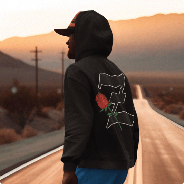 Person wearing a black hoodie with a red rose design and black Z logo, skateboarding on a road into the sunset.