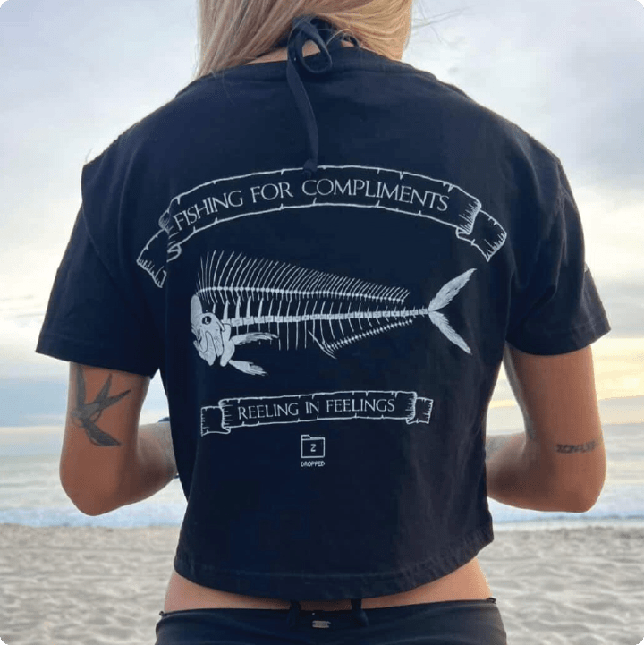 Young woman wearing a black crop top shirt with a fish skeleton design and text on the beach.