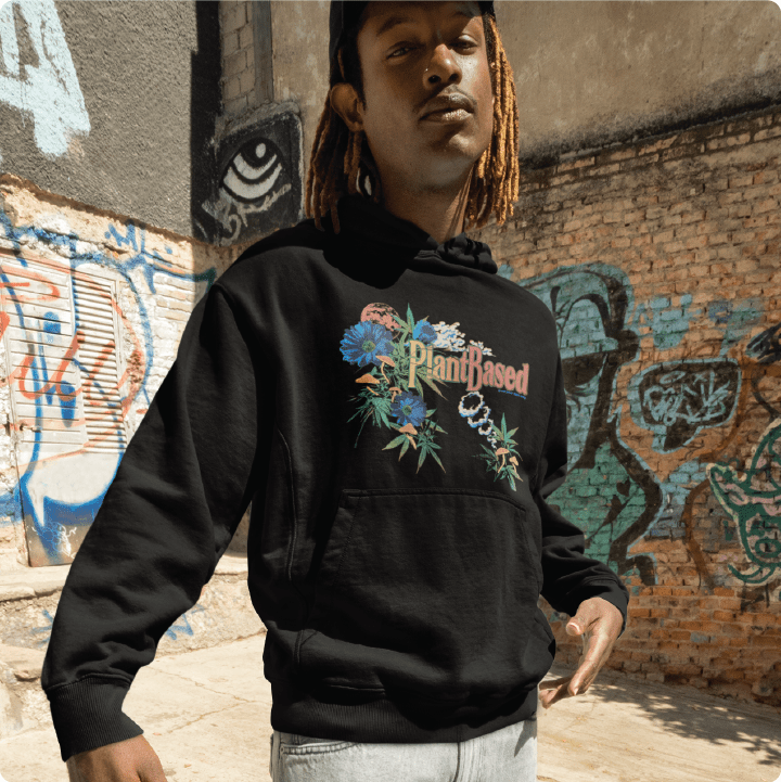 Person wearing a unisex black hoodie with 'Plant Based' text and cannabis floral designs standing in front of a graffiti-covered wall.