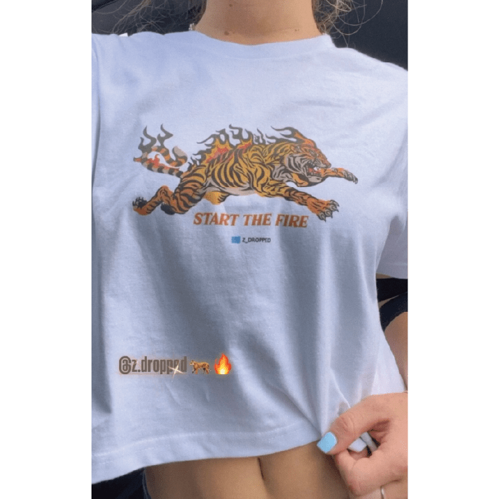 Young woman taking a selfie wearing a white crop top t-shirt with a tiger graphic and 'Start The Fire' text.
