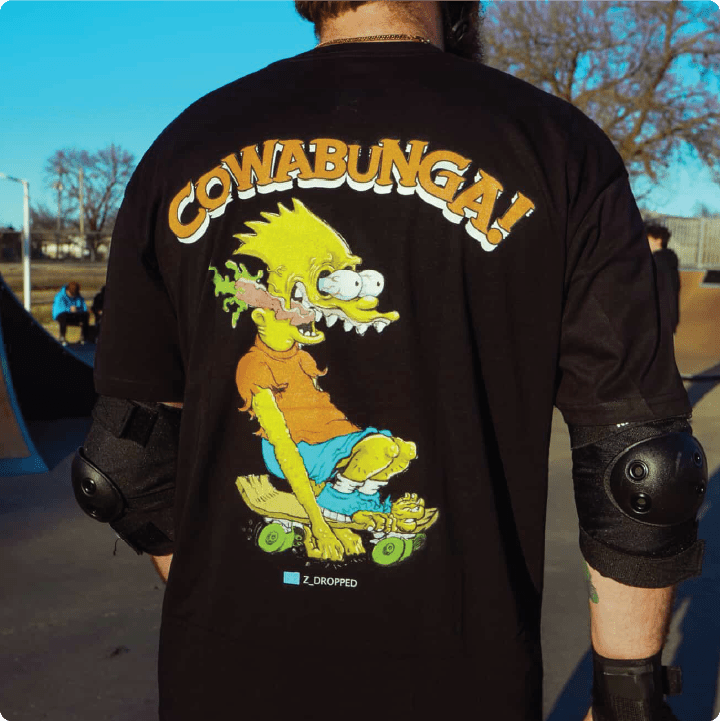Rear view of a man wearing a black t-shirt with a bart roth cartoon character and 'Cowabunga!' text at a skate park.
