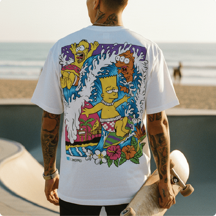 Rear view of a  man standing at a beach skatepark wearing a white bartsunami t-shirt holding a skateboard.