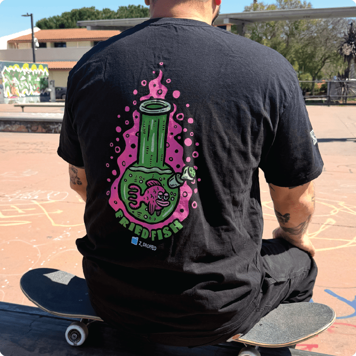 Rear view of a black shirt with a neon green bong graphic and pink fish inside, surrounded by pink bubbles and “FRIED FISH” text at a skate park.