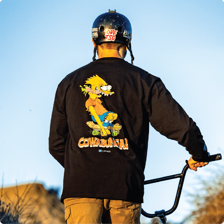Rear view of a bmx rider wearing a black long-sleeve shirt with a Ed Roth Bart Simpson character and 'Cowabunga!' text oitdoors.