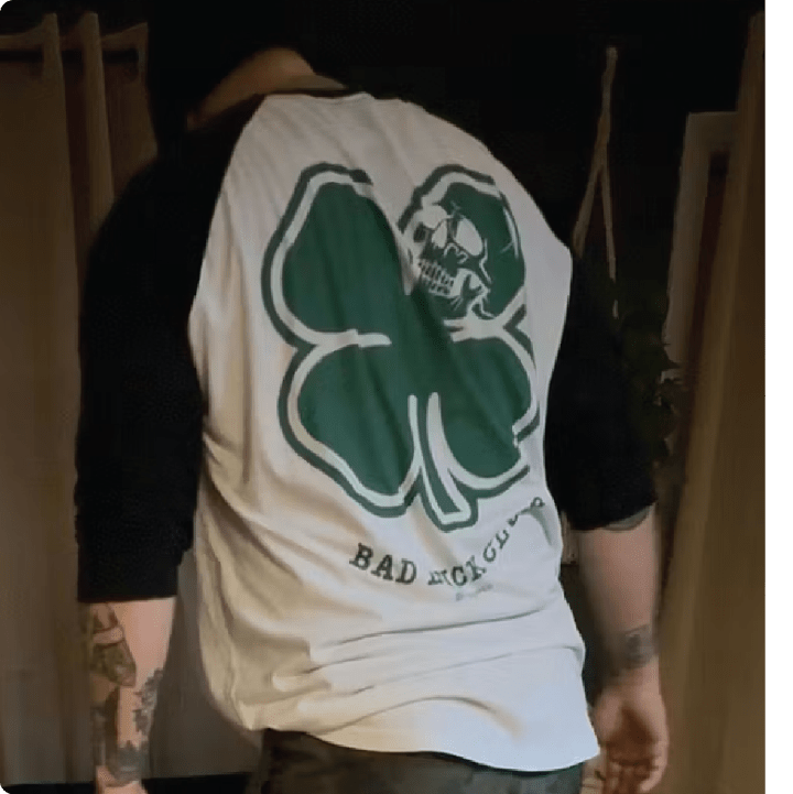 Rear view of a person wearing a white 3/4 sleeve raglan with a green clover and skull design, text 'Bad Luck Club' on the back.