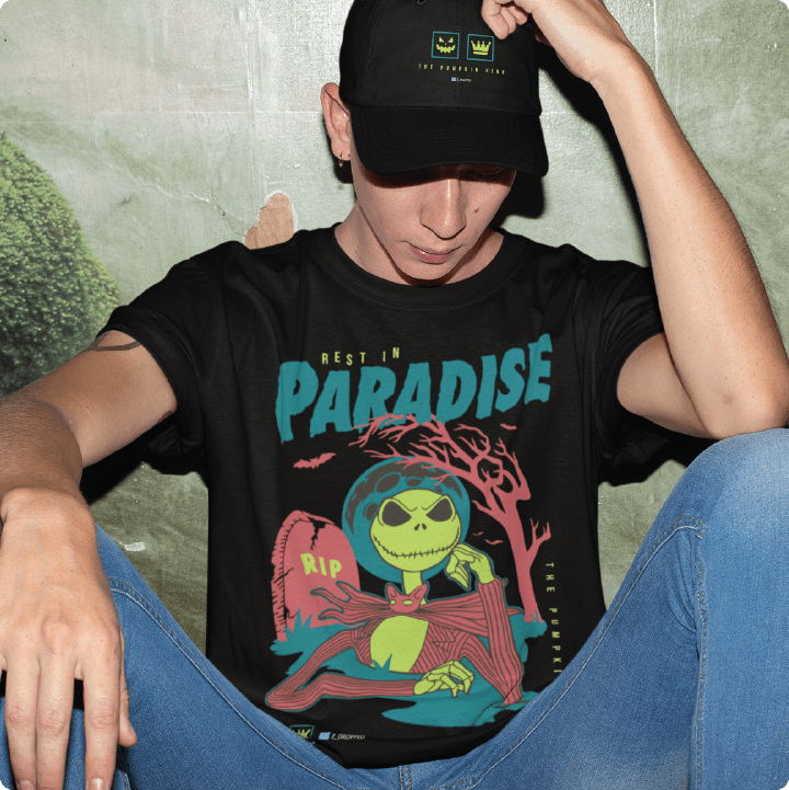Person wearing a black zdropped jack-o-lantern t-shirt with a colorful graphic design and text, sitting against a textured wall.