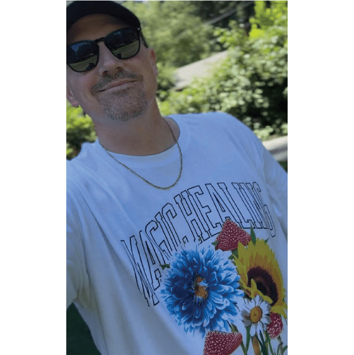 Front view of a man wearing a white t-shirt with floral designs and 'Magic Healing' text outdoors taking a selfie.