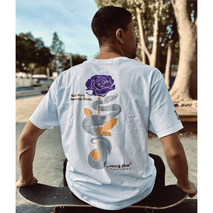 Rear view of pro skateboarder nick tucker wearing a white mamba quote t-shirt with a snake and rose design, sitting on a skateboard outdoors.
