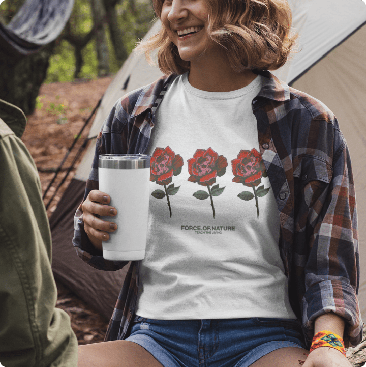 Person wearing a white t-shirt with red floral design and plaid shirt, holding a white tumbler outdoors.