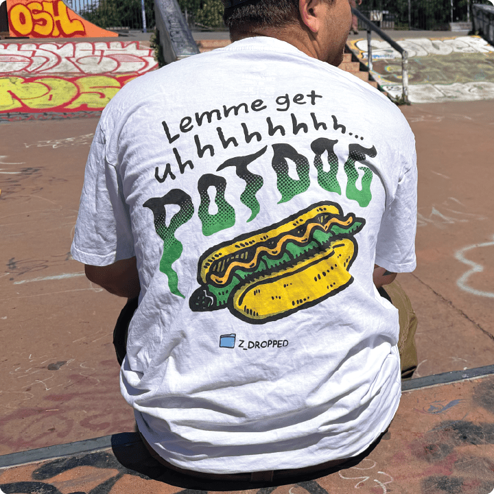 Rear view of a white shirt with a green blunt hot dog with green smoke and black “Lemme get uhhhhhh… POTDOG” text at a skate park.