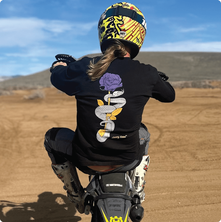 Person on a dirt bike wearing a black mamba quote long sleeve tee with snake and lakers rose design in an outdoor setting.