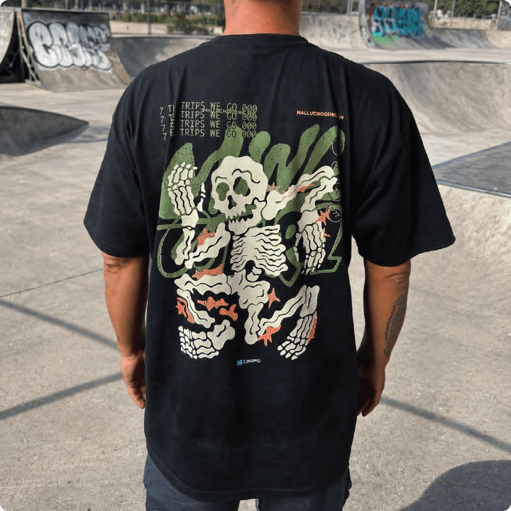 A person standing at a skatepark wearing a black t-shirt with a large green, white, and orange graphic skeleton print on the back. The background shows concrete ramps and graffiti.