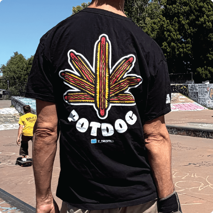 Rear view of a black shirt with a weed leaf made of hotdogs and white “POTDOG” text at a skate park.
