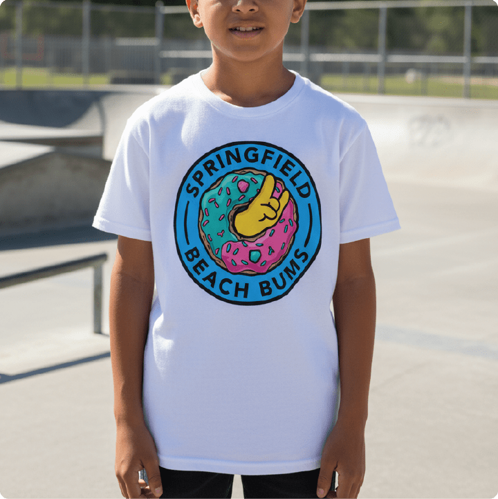 A child wearing a white short-sleeve t-shirt with a colorful circular graphic on the chest that reads 'SPRINGFIELD BEACH BUMS' around a donut illustration, photographed outdoors at a skatepark.