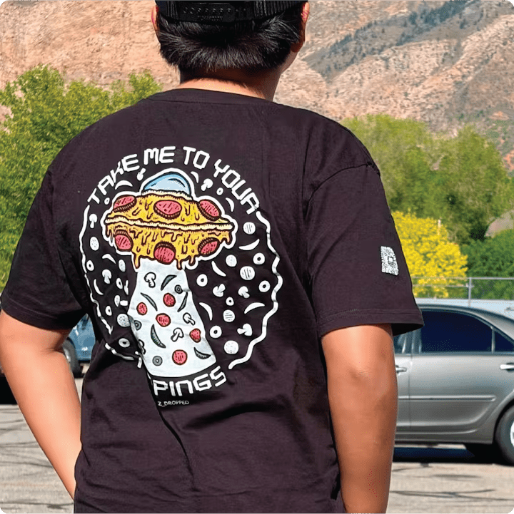 Rear view of a youth Tee with a pizzabduction UFO and “Take Me To Your Toppings” text standing outdoors.