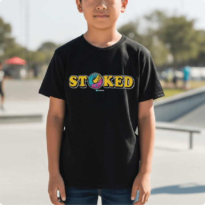 A child wearing a black short-sleeve t-shirt with a colorful 'STOKED' graphic across the chest, standing outdoors in a skatepark setting.